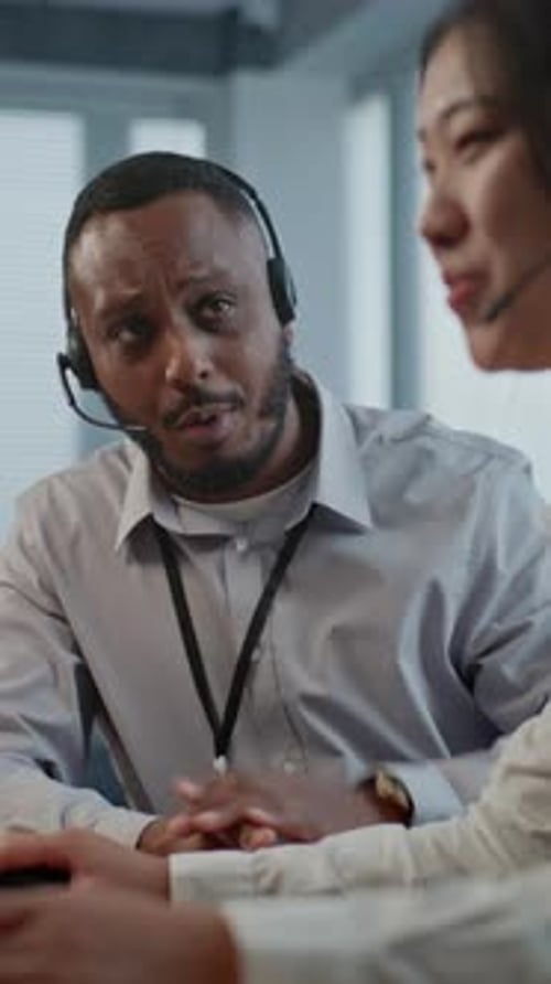 Two Multiethnic Hotline Operators in Headsets Use Computer Having Conversation with Client