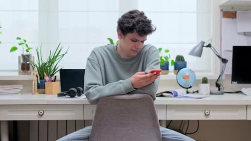 Hübscher junger Mann sitzt mit Smartphone auf einem Stuhl zu Hause