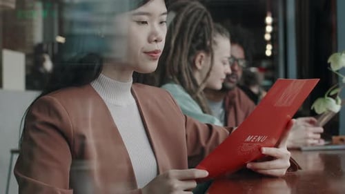 Asian Woman Sitting at Wooden Counter in Restaurant, Carefully Reading Menu