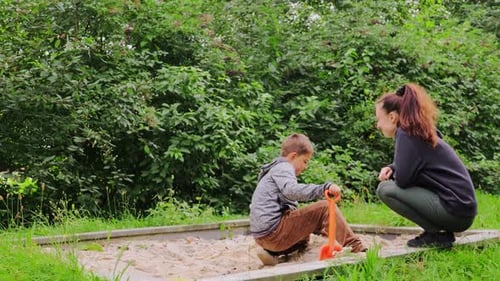 A young mother or nanny engages with a child in a playground filled with lush greenery.
