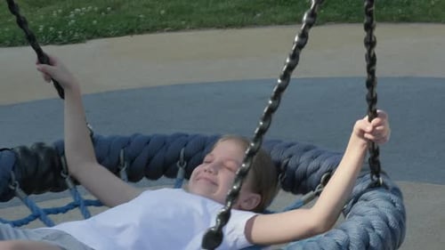 Little Girl Smiling Swinging on Blue Round Swing in Playground on Summer Day