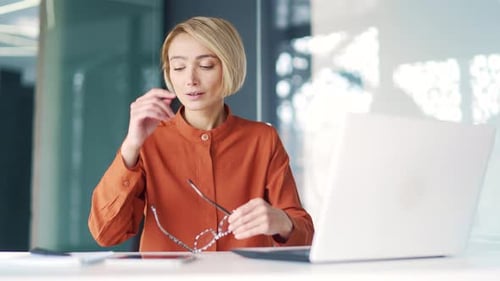 Überarbeitete junge Frau mit Kopfschmerzen, müde, am Laptop zu arbeiten, während sie am Arbeitsplatz am Schreibtisch in