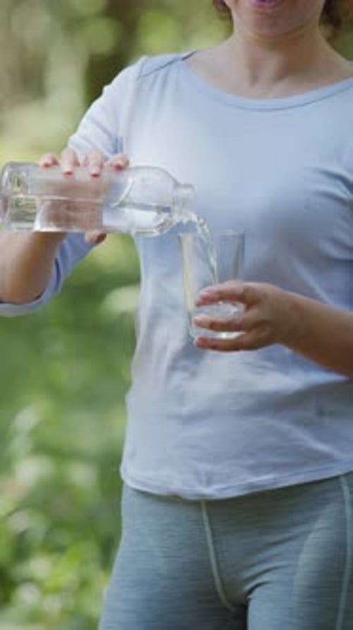 Woman is pouring pure water into a glass from a bottle, a blurred green nature background.