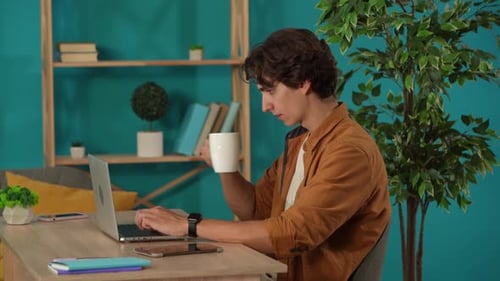 Medium Shot Capturing a Man Working on a Laptop in His Home Office and Drinking Coffee Young Male