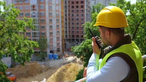 Männlicher Vorarbeiter mit Schutzhelm steht auf der Baustelle und gibt Anweisungen per Walkie-Talkie