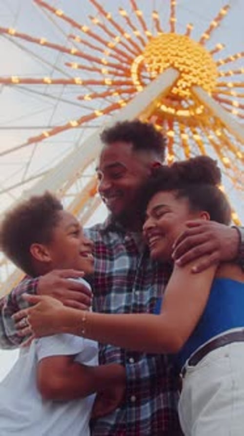 Optimistic Couple Hugging Son on Background of Ferris Wheel