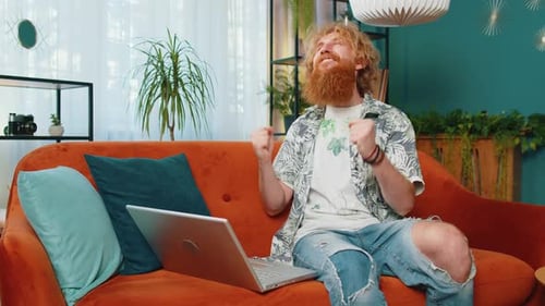 Man Celebrates Success Using Laptop on Sofa