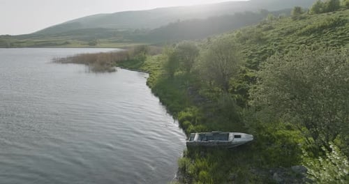An abandoned motorboat on the shoreline of a calm lake in the early morning.