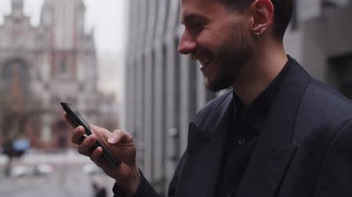 Smiling Young Man Standing on the Background of the City Uses a Smartphone A Bearded Man in a