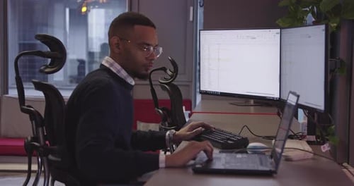 Young African American with Glasses Sitting in a Modern Coworking Office and Working at a Computer