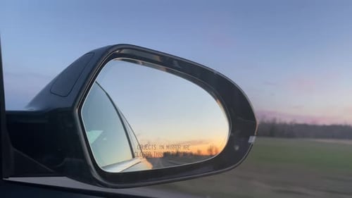 Reflection in Car Mirror while Driving during sunset on long empty road in rural area - late evening