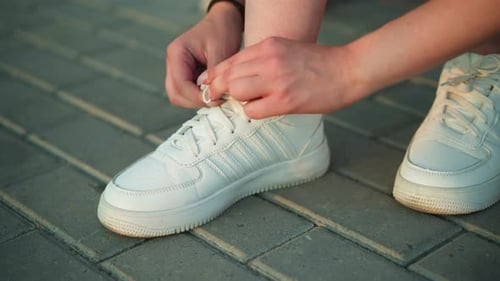 CloseUp of Person Tying Shoelaces of White Sneakers on Pavement Path Outdoors