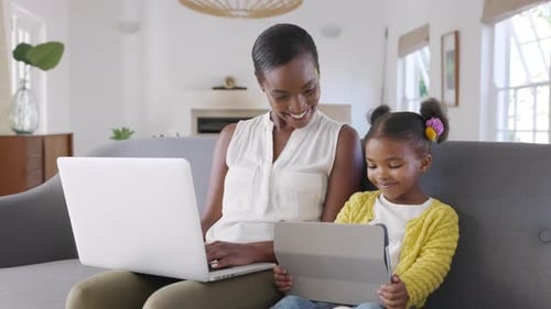 Smiling african mother works on laptop at home while daughter uses digital tablet