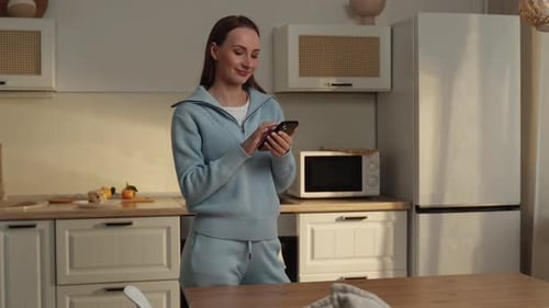 Young Happy Housewife Dressed in a Blue Sweater Holding a Mobile Phone in Her Hands Looking for a