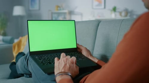 Man Lying on His Sofa At Home, Uses Laptop with Green Mock-up Screen. Over The Shoulder Shot of Ano