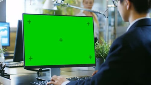 In the Office Businessman works on a Desktop Personal Computer with Mock-up Green Screen. Colleague