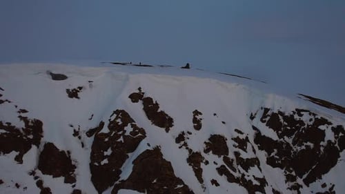 Aerial flyover shot of two people standing on the snowy peak of a mountain on a blue cloudy sky with
