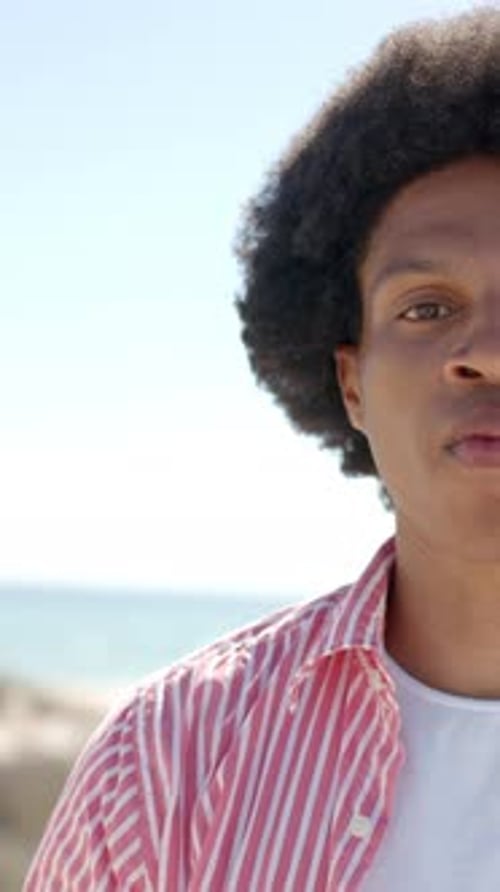 Vertical video: Smiling African American man enjoying sunny day at beach, wearing striped shirt