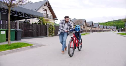 Happy Young Father Teaching Small Cute Smiled Son in Helmet to Ride a Bike at Street Near House