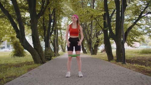 Athletic Fit Sport Girl Doing Squats Using Fitness Elastic Rubber Band Workout in Park at Morning