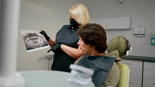 Dental Office Blonde Dentist in a Black Uniform Consulting Patient Showing a Photo of the Teeth