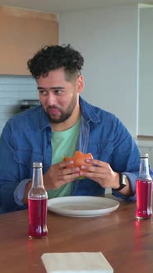 Vertical video: Hunger driving man in denim taking bites at kitchen counter with hamburger, bottles