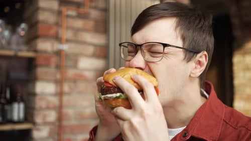 Male Customer in Glasses Red Shirt Eating Burger at Restaurant Brick Wall Background Blurring