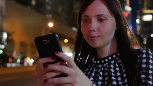 A Young Woman Using a Smartphone at Night in an Urban Area Illuminated By City Lights