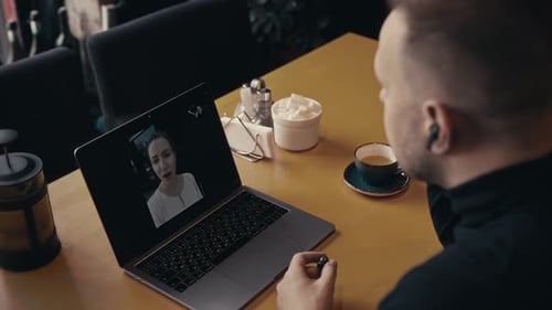 Ein Mann in einem dunklen Pullover sitzt mit einem Laptop in einem Café und chattet per Video mit einer Frau. Ein Videoanruf
