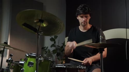 A Man Dressed in a Black Tshirt and Shorts Sits at a Drum Kit in a Home Studio He Plays the Drums