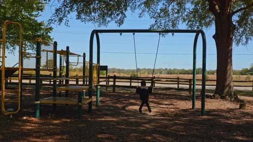 A young child with her back turned swinging on a swing on a playground with a road in the background