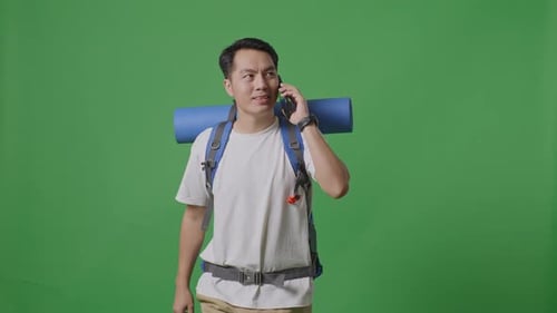 Asian Male Hiker Talking On Smartphone And Looking Around While Walking On Green Screen