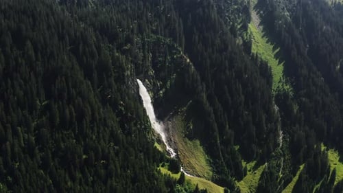 Aerial wide shot of majestic waterfall cascading through dense alpine forest on a sunny day. Water c