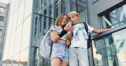 Two teenage students boy and girl friends using smartphone exchange lectures near school building