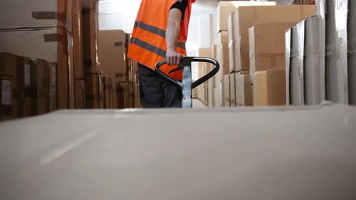 View from pallet truck. Young male worker in uniform is in the warehouse pushing boxes