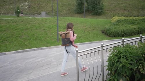 Young College Student Walking Through Green Urban Park Carrying Backpack and Moving Toward Morning