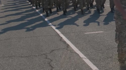 Soldiers marching down city streets, wearing camouflage uniforms. Low angle view of their legs and f