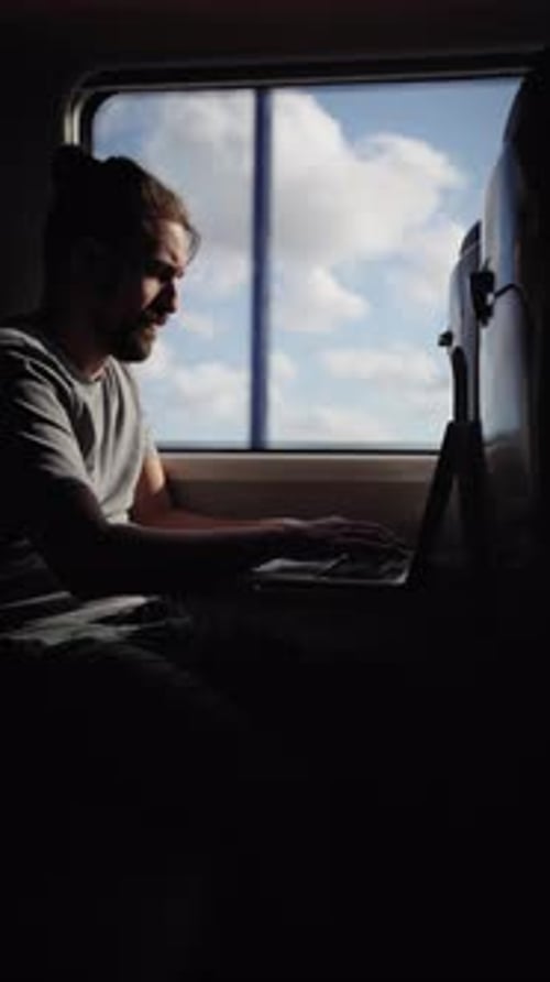 Junger kaukasischer Mann sitzt im Zug und tippt und schaut durch das Fenster auf Laptop-Wolken
