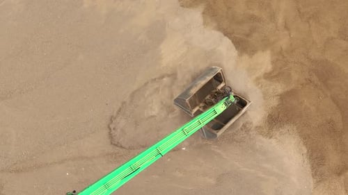A top-down aerial view of an excavator's green arm and scoop digging into the sand, creating a dust