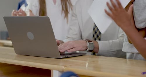 Close view follows male adult teacher using laptop while young multicultural student group passes