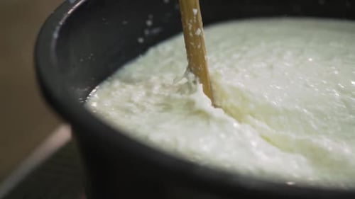 Cheese and Curd production process. Fermented milk quickly mixed in black bucket with a wooden ladle
