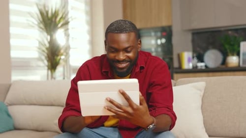 Man Sitting on Couch Uses Tablet Device at Home