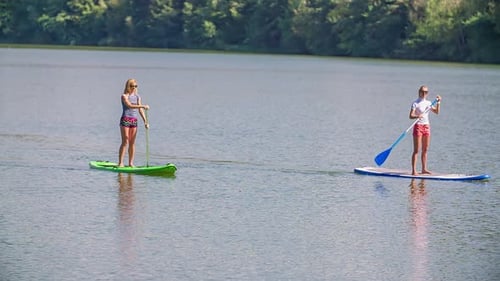 Two blonde girls on stand up paddle boards (SUP) paddling down stream on a river in the sunshine, ha