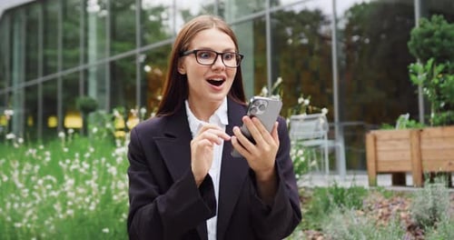 Portrait of Young Caucasian Woman in an Urban Park Holding a Smartphone and Reacting with Surprise