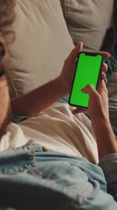 Young man lying on sofa uses mobile phone with green mock-up screen chroma key