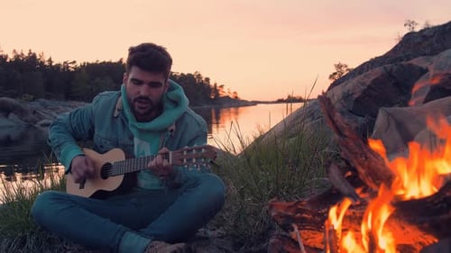 Jeune beau musicien jouant de la guitare et chantant devant un feu de joie sur une île au bord de l'océan.
M