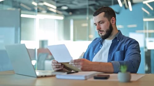 Man Reads Document at Office Desk with Laptop