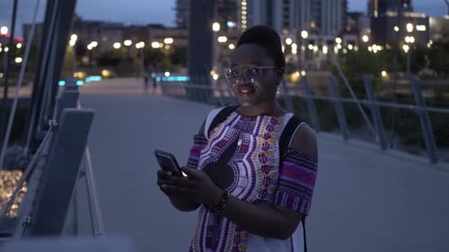 Happy young woman uses smart phone on city footbridge at night