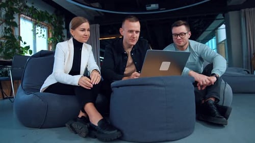 Office staff sitting close on bean bag chairs look at the laptop in front of them.