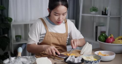 Asian woman enjoying making fresh fruit whipped cream sandwich on table at home.